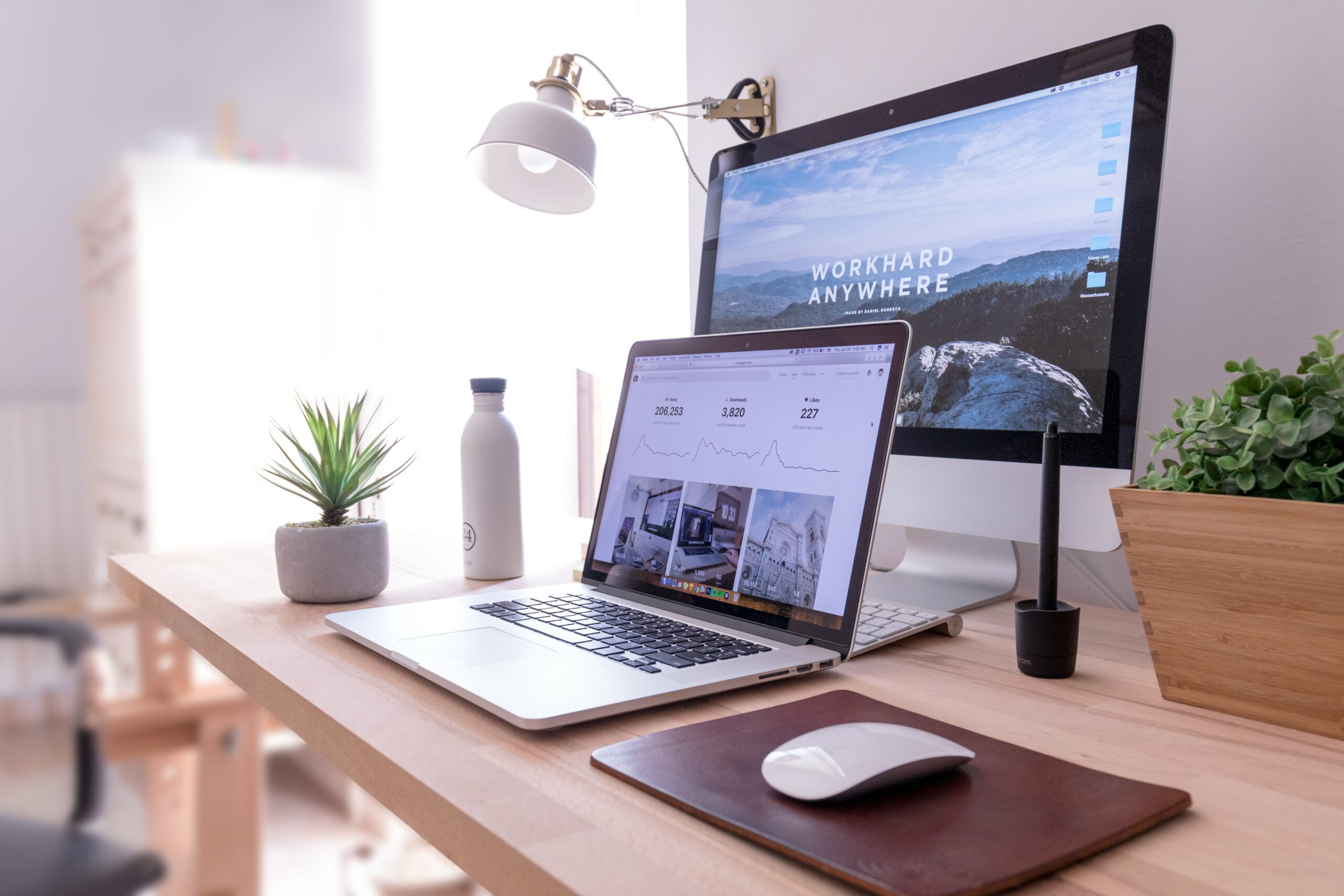 A laptop sitting on a desk in front of another computer monitor displaying a large background image. Two plants site in the background and a mouse sits on the desk in front of the laptop on a brown mousepad