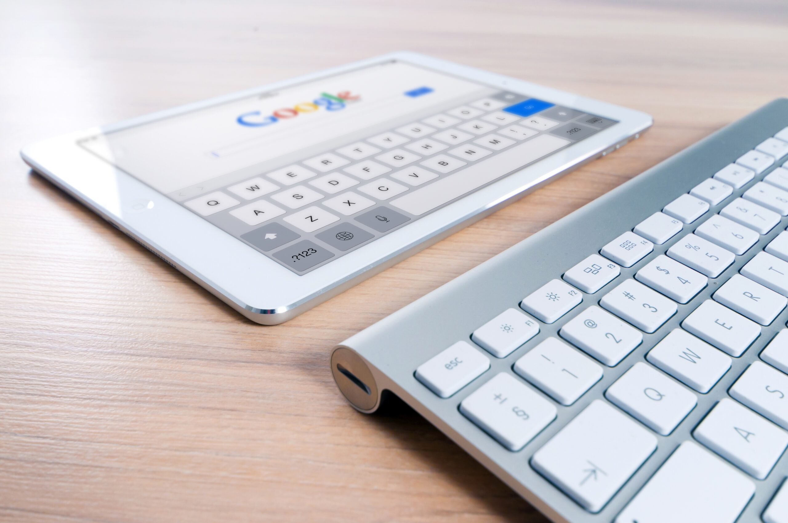 White tablet on a wooden table open to Google's homescreen with a keyboard in front of it