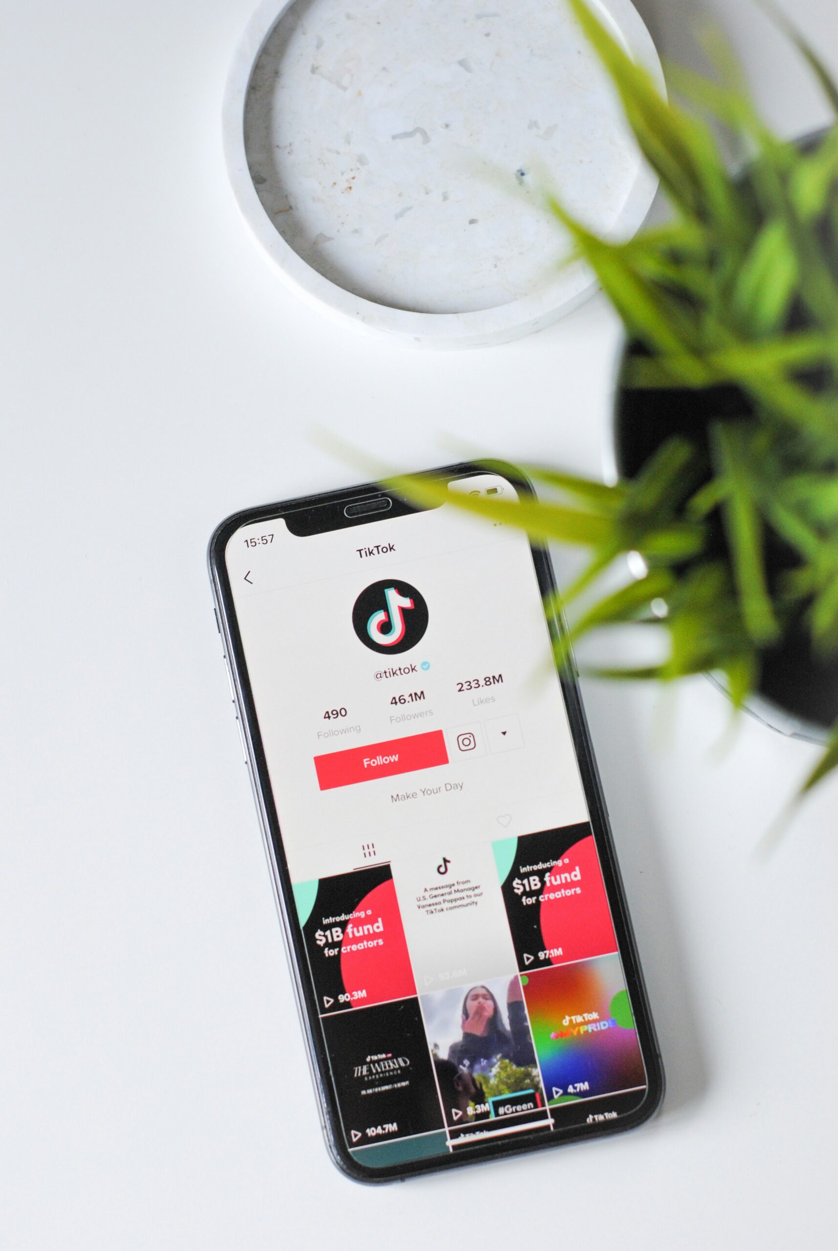 A cell phone on a white table with a plant next to it. The cell phone displays a basic TikTok account feed.